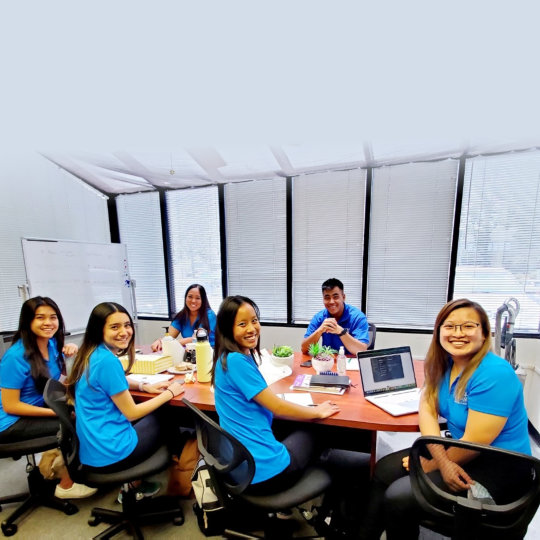 A group of people wearing blue shirts sitting around a conference table with notepads, laptops, and a whiteboard in the background.